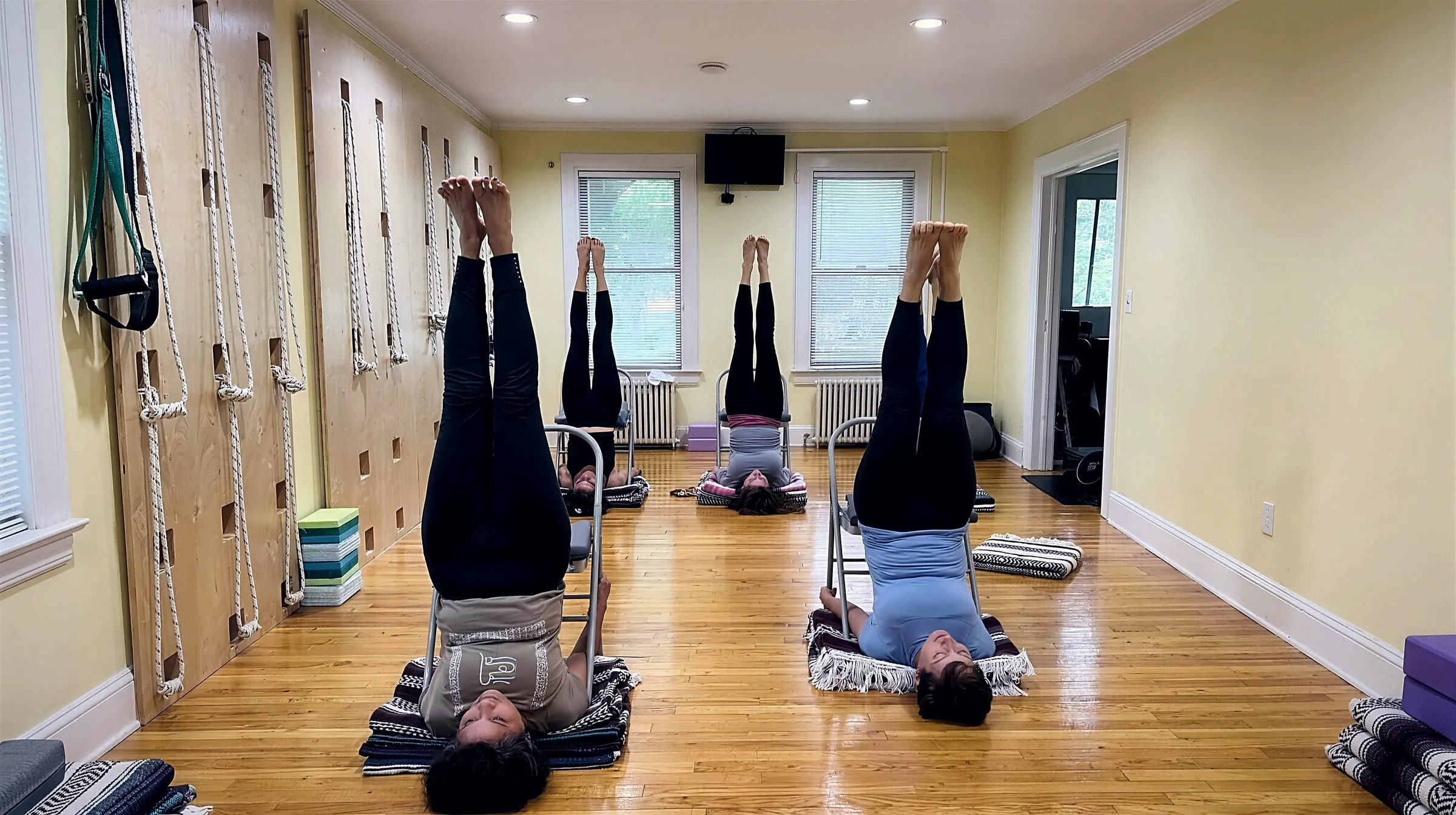 Four members in a supported shoulderstand over folding chairs, blankets under their shoulders, beside the studio’s rope wall