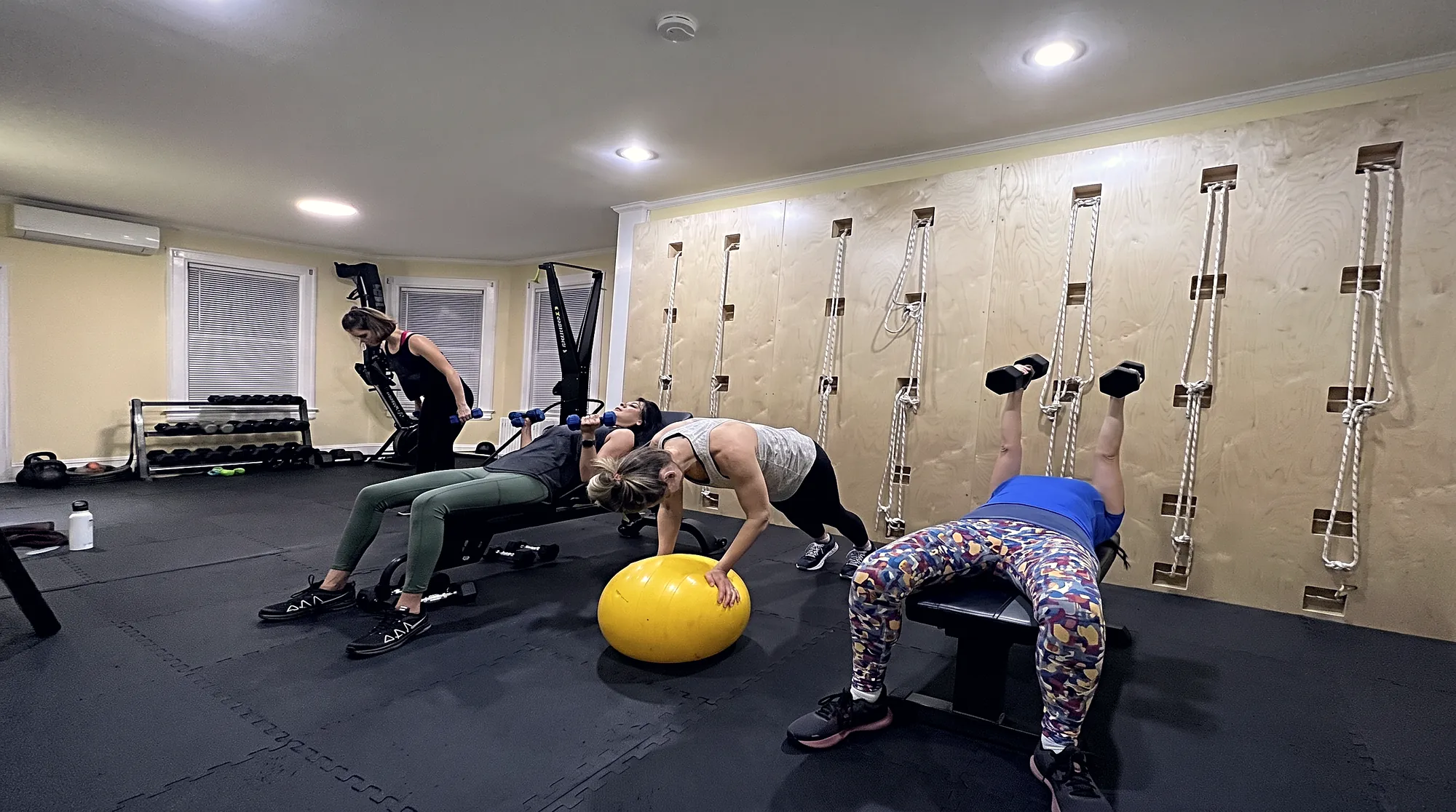 A coach talking a client through a kettlebell squat, a rack of kettlebells and dumbbells on the shelves behind them